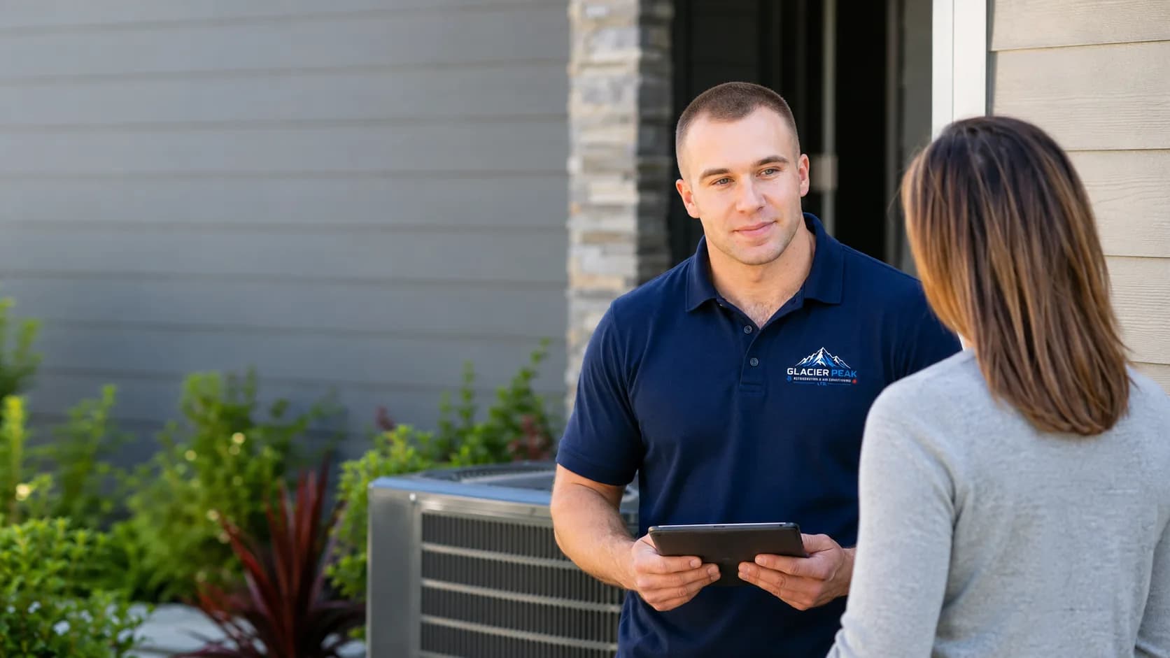 HVAC technician with a tablet talking to a homeowner beside an outdoor condensing unit at a house